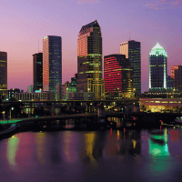 Sunset view of a vibrant city skyline reflecting on the water, with illuminated skyscrapers in shades of pink, purple, and green.