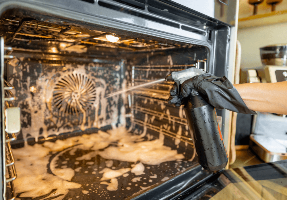 A person in black gloves sprays foaming cleaner inside a dirty oven, with bubbles and grime visible on the surface.