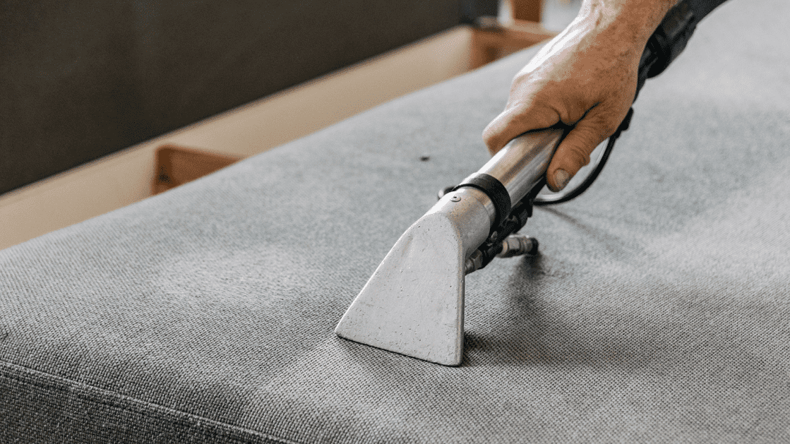 A person is using a vacuum cleaner attachment to clean a gray upholstered surface.