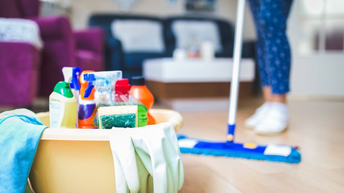 A cleaning scene featuring a basket with cleaning supplies, including sprays and sponges, and a person using a mop on the floor.