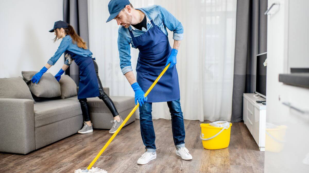 Two people cleaning a living room; one is mopping the floor while the other adjusts cushions on a couch. They are wearing blue aprons and gloves. A yellow bucket is nearby.