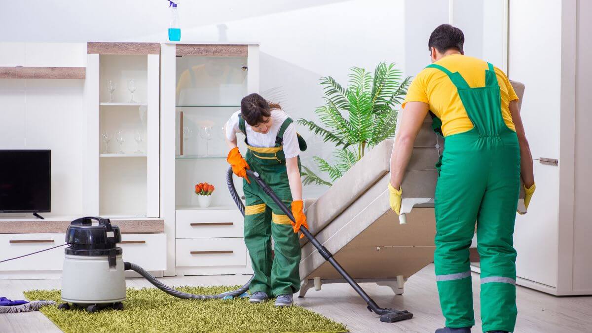 Two people in work uniforms are cleaning a living room. One person is using a vacuum cleaner while the other is lifting a sofa.